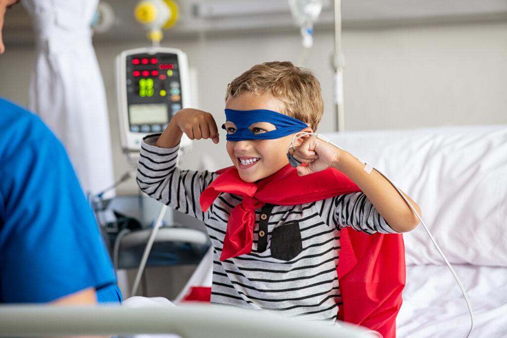 Cheerful little boy in a hospital bed dressed as a superhero.