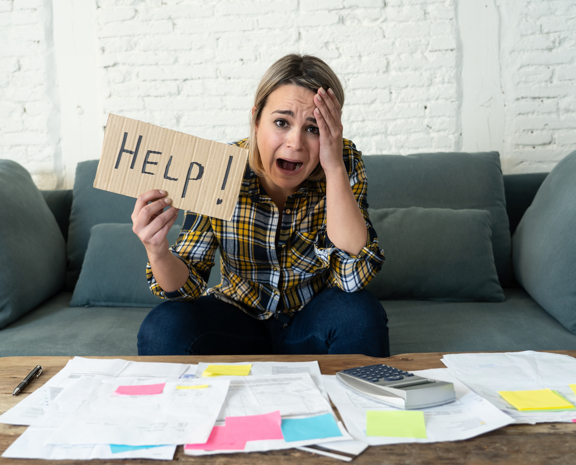 Portrait of worried desperate young woman showing Help sign in front of a pile of medical bills.